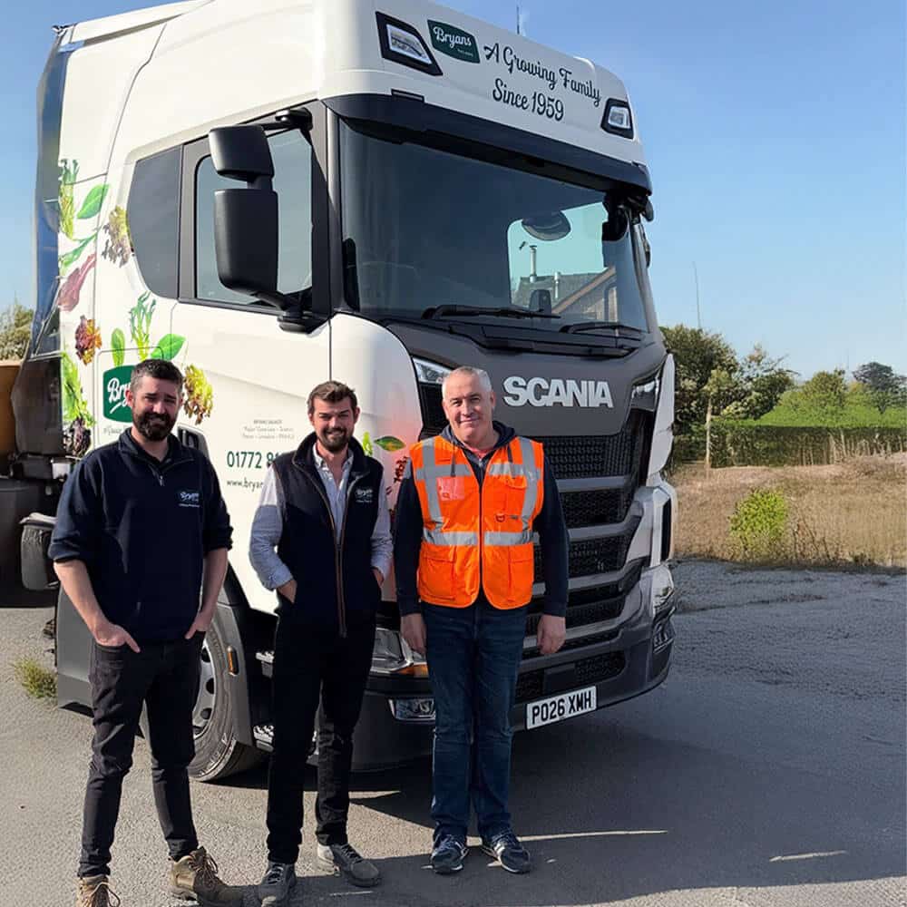 Three men standing proudly in front of a white Scania S500 tractor unit featuring custom Bryan Salads branding. The truck displays a "Bryans Salads: A Growing Family Since 1959" decal on the sun visor and vibrant fresh produce graphics on the side of the cab. One man wears a high-visibility orange safety vest, while the others are in branded casual workwear, all positioned against a clear blue sky and rural background.