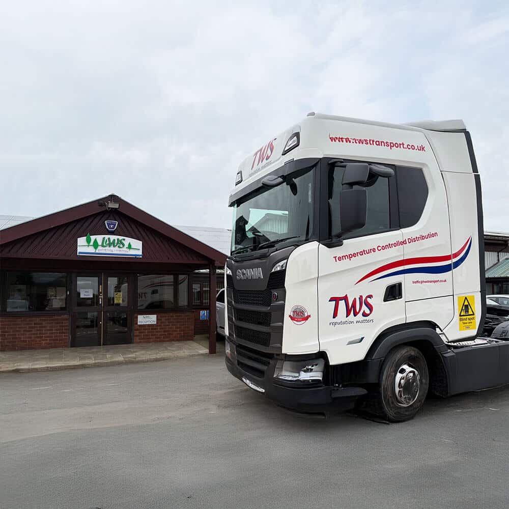 A side-angle shot of a new white Scania S500 tractor unit parked in front of the LWS reception building. The vehicle features the red and blue TWS "reputation matters" logo, the "Temperature Controlled Distribution" text, and the company website URL on the air deflector. The LWS group logo is visible on the brown wooden building in the background under a cloudy sky.