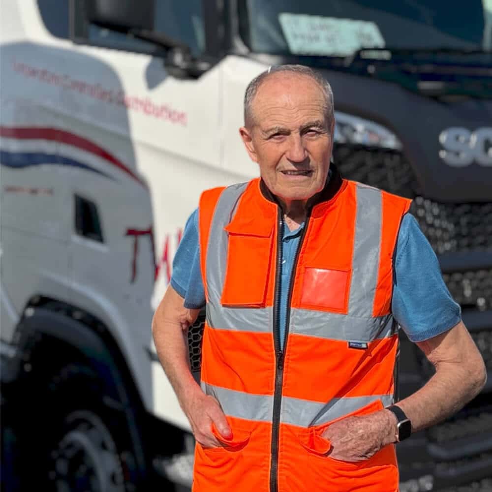 A photograph of a smiling elderly man, wearing an orange high-visibility safety vest over a blue polo shirt, with his hands in his pockets. He is standing in front of a white Scania tractor unit, which has visible, though blurred, 'TWS' and 'Temperature Controlled Distribution' branding on the side, set within a truck yard on a sunny day.